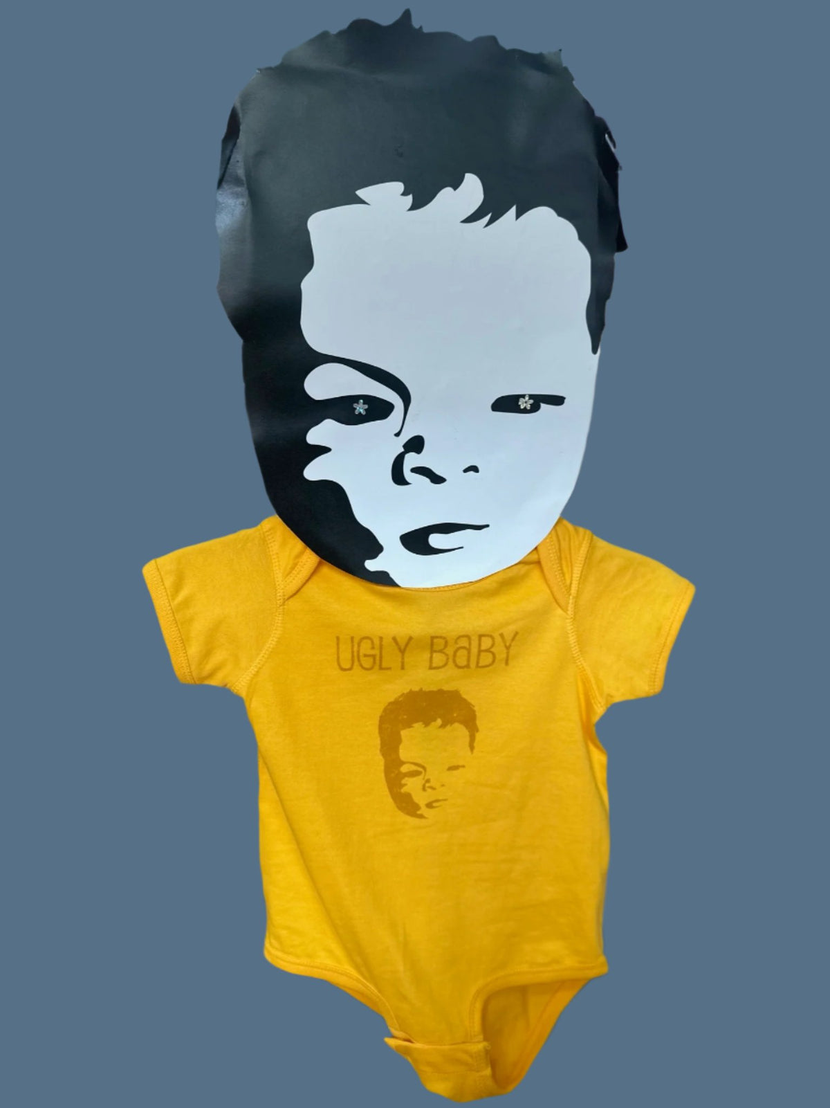 Gold colored infant bodysuit with the Ugly Baby logo printed in darker gold ink. The rack that the bodysuit is hanging on has a large, oversized head in the shape of the Ugly Baby logo at the top. 