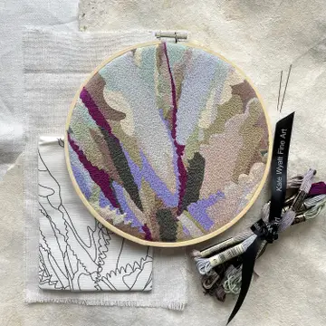 An example of the finished embroidery kit agave with blue, purple and natural colors. A cloth with the printed pattern is folded below the example. 