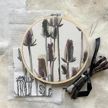 An example of the finished embroidery kit - thistle with blue, purple and natural colors. A cloth with the printed pattern is folded below the example. A batch of embroidery floss is tied with a ribbon and sitting on the table next to it. 