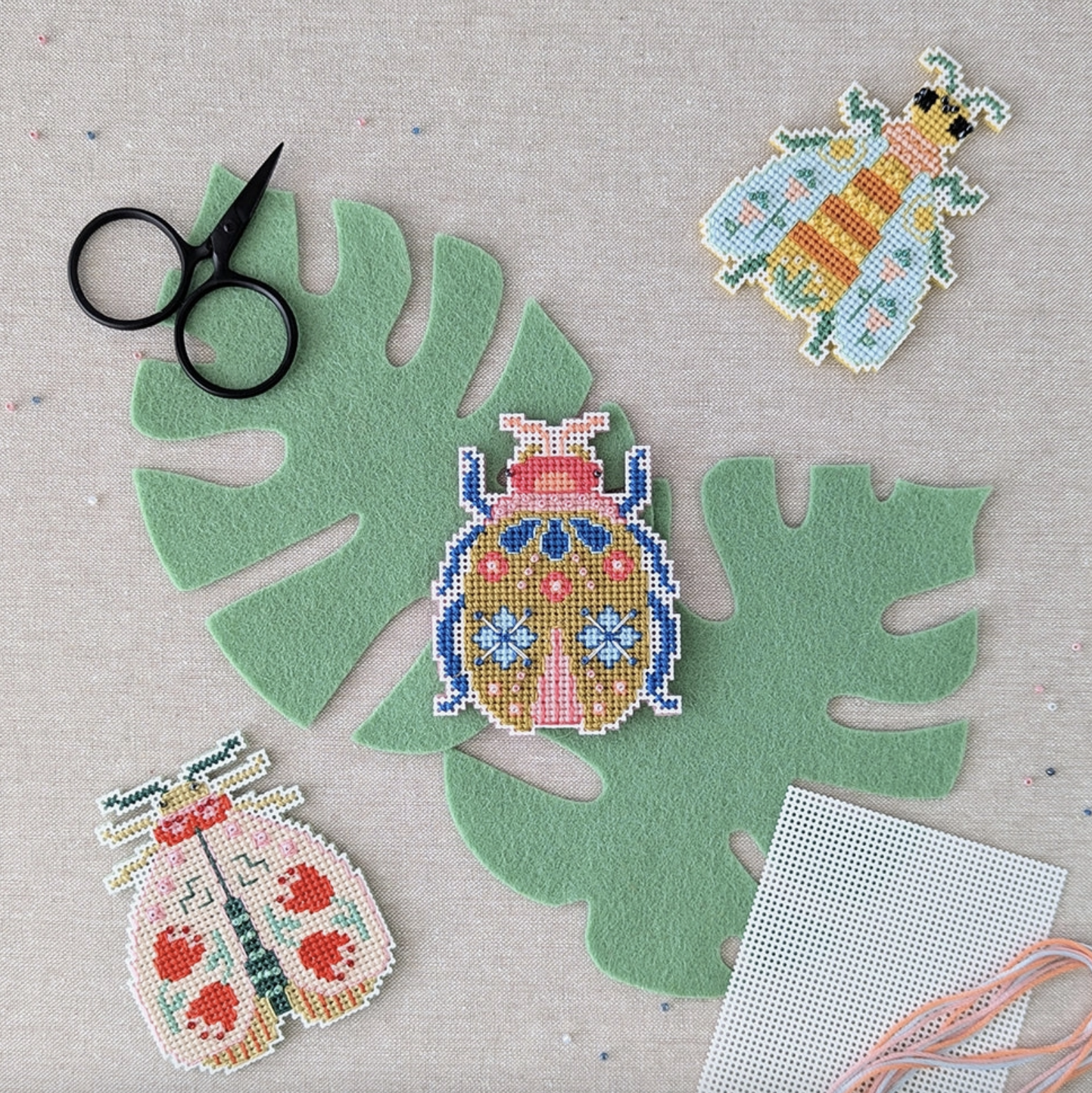 All three of the Blooming Bugs kits laid out on a table: bee, beetle and moth. They are laying on top of a felt monstera leaf and there are sewing supplies scattered around the background: cute little black embroidery scissors, perforated paper and embroidery floss. 