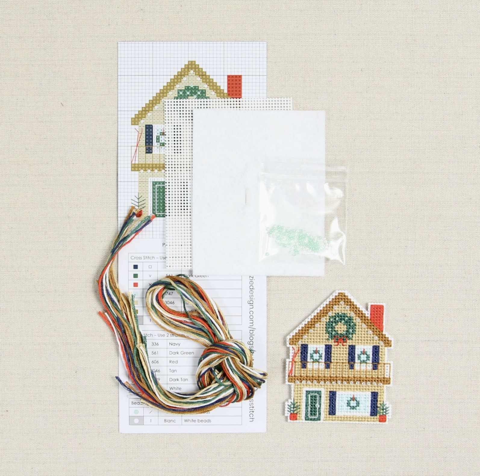 All the items included in the holday home chalet cross stitch kit laid out on a table: pattern, embroidery floss, perforated papey, needles and a tiny bag of beads. 