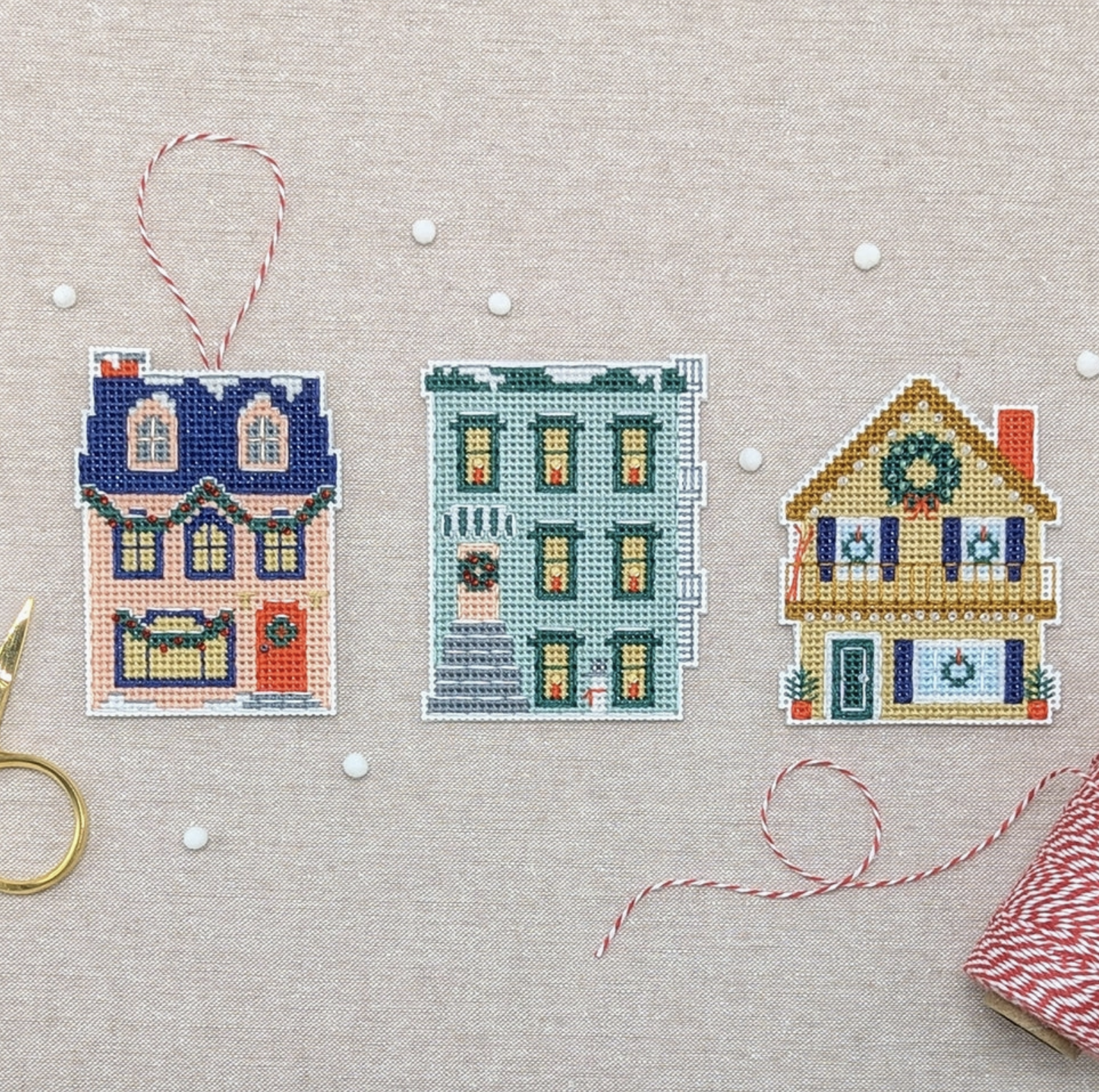 A photo of all three of the holiday homes cross stitch patterns laid out on a table with white beads scattered around them. 