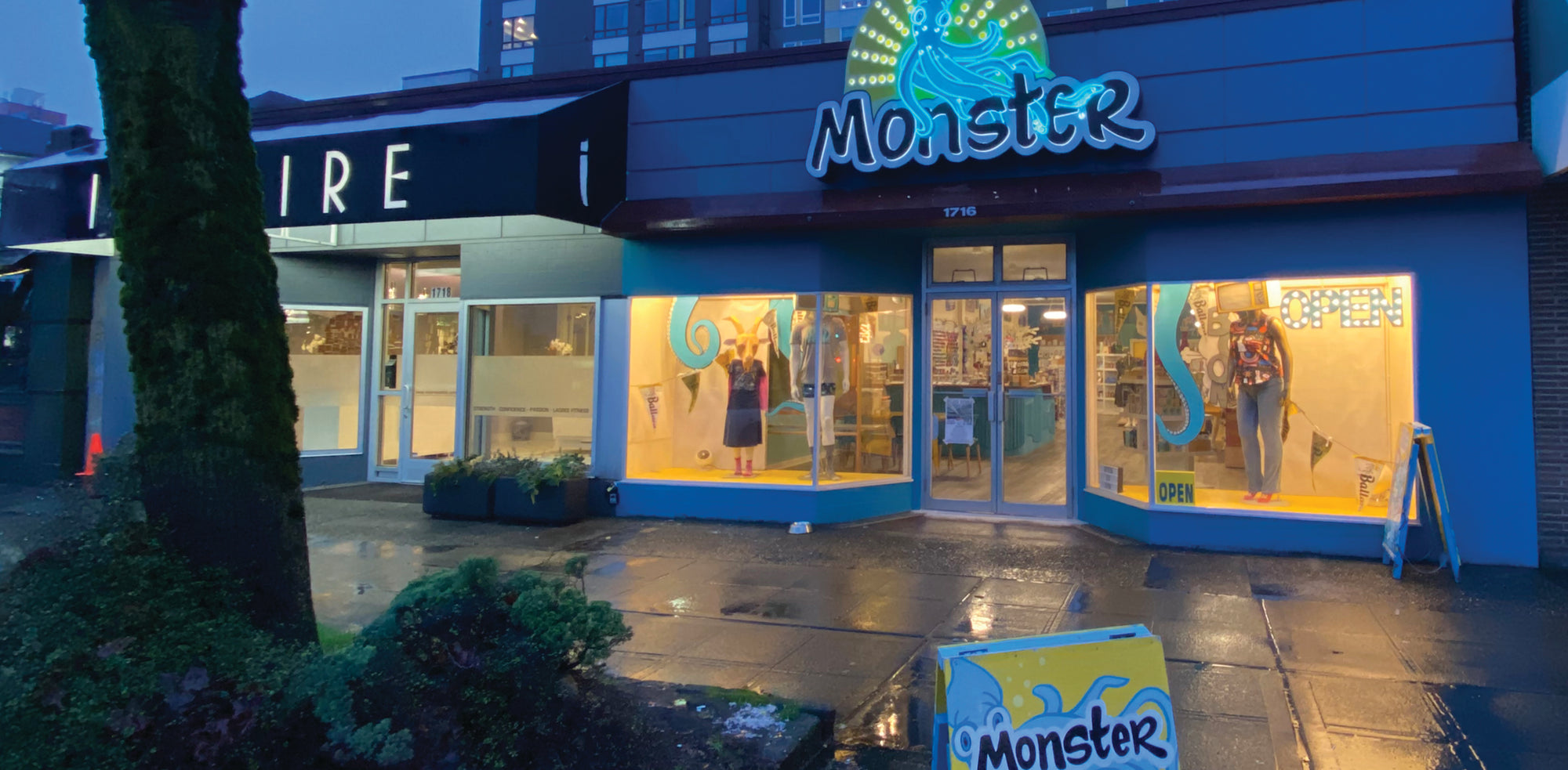 Exterior of Monster at dusk. The neon sign says "Monster" and has a friendly blue squid with sun beams behind it. The storefront is painted blue and tentacles reach down through the ceiling. 
