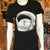 Black crew neck t-shirt with the image of a white cat snuggled up in an astronaut helmet. There are stars in the background.