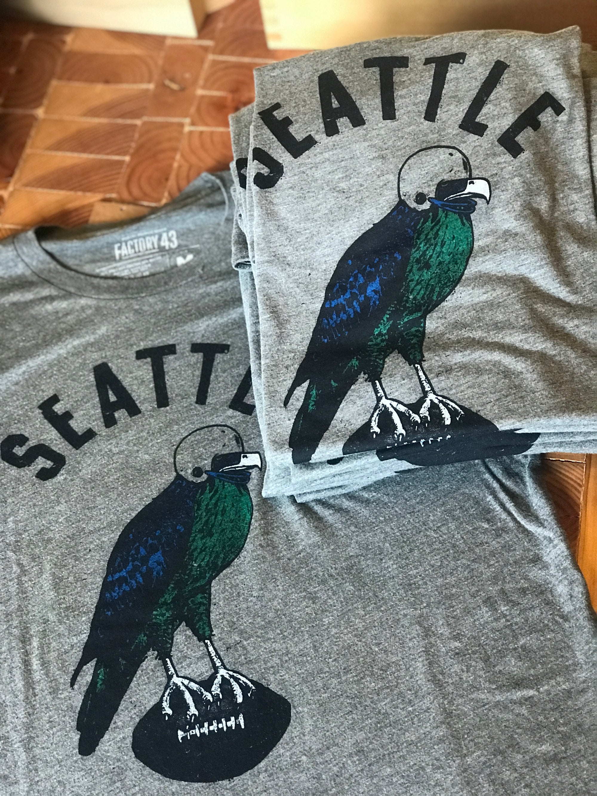 A stack of folded t-shirts that say Seattle and have a hawk carrying a football depicted on them. 
