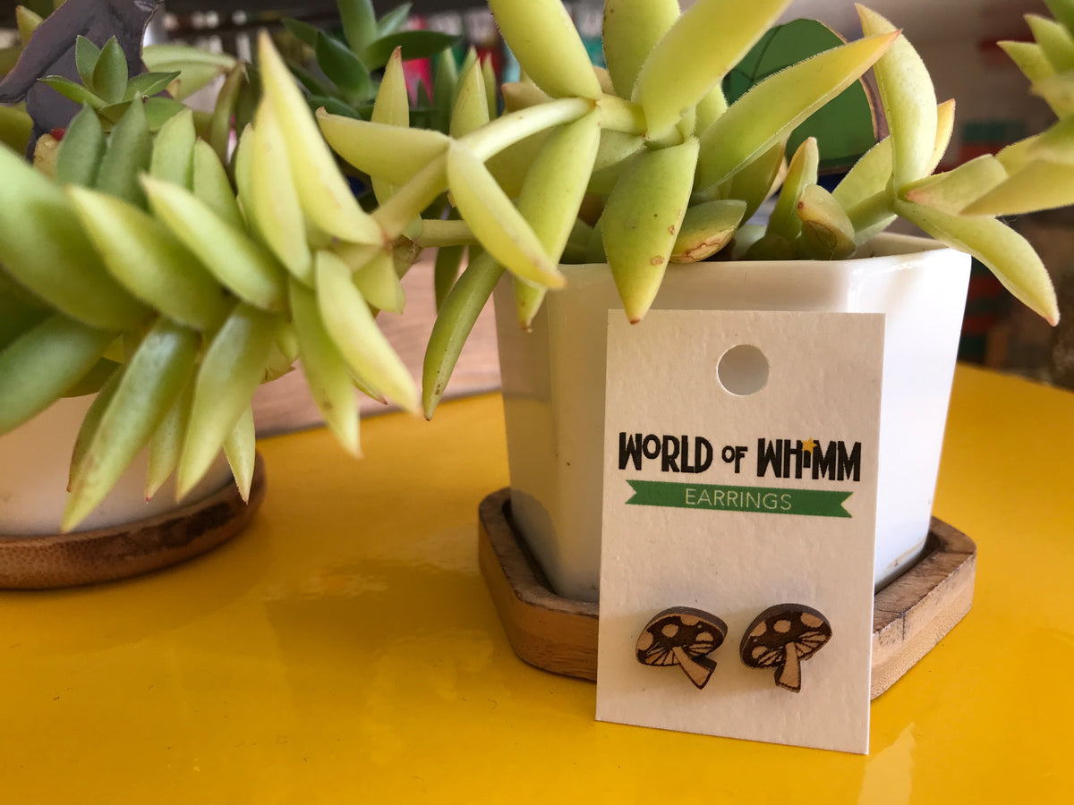 Wee laser cute earrings in the shape of mushrooms are attached to a white backing card that says &quot;World of Whimm.&quot; They are leaning against a small succulent plant on a bright yellow table. 
