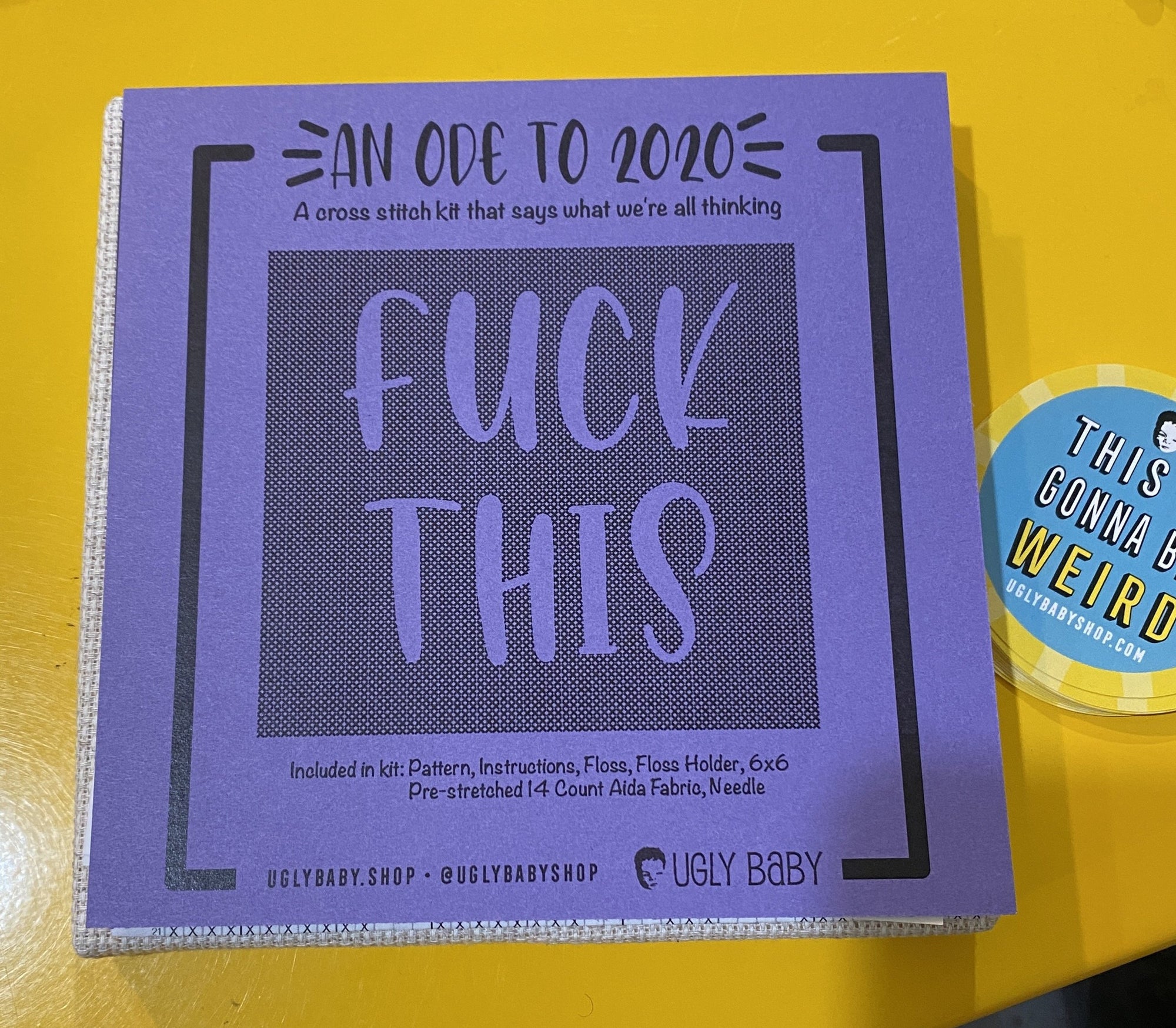A cross stitch kit. The packaging is purple and says "An ode to 2020. Fuck this." The packaging is dark purple against a bright yellow background. There is a sticker in the right corner that says, "This is gonna be weird." 