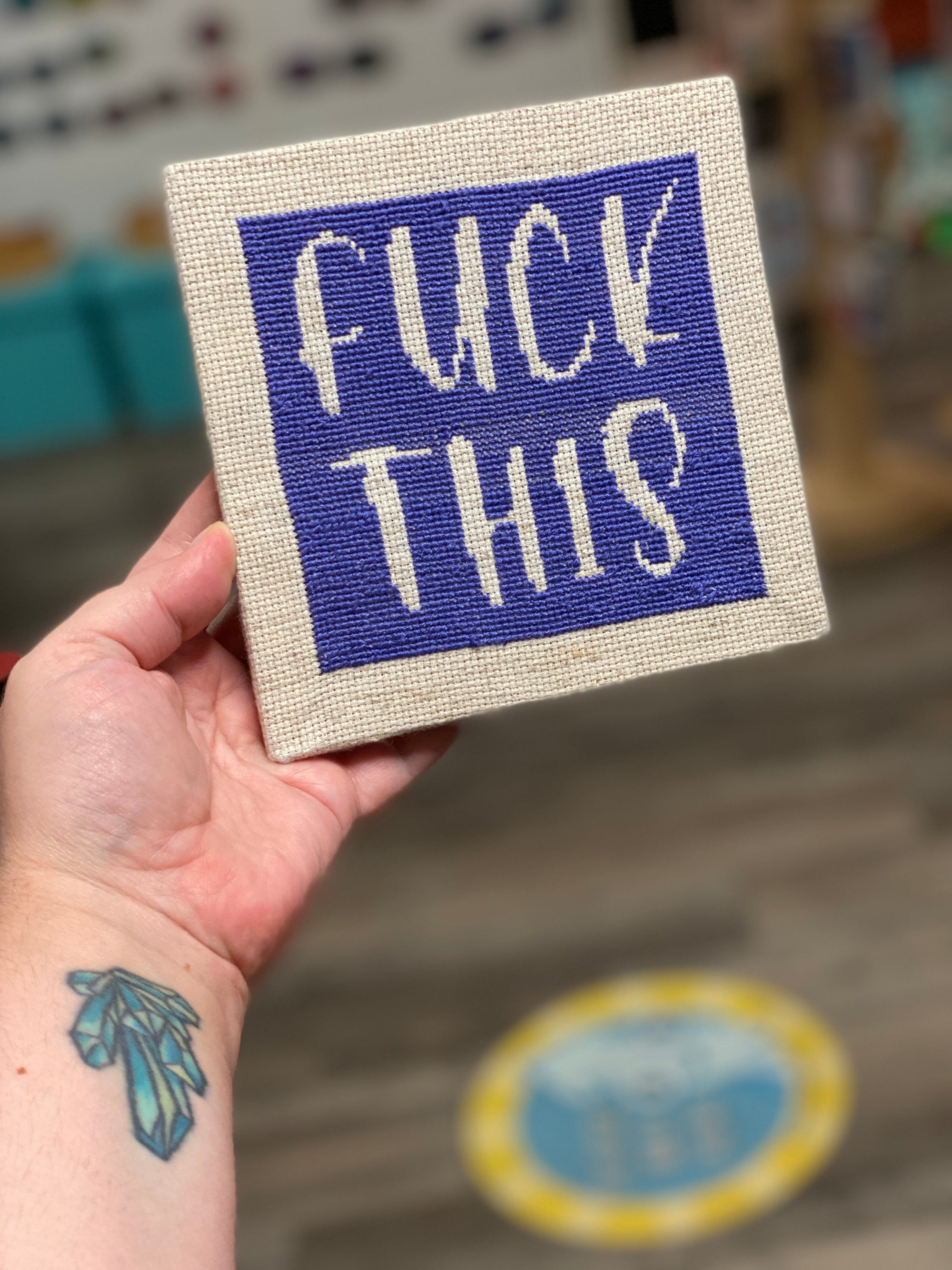 A cross stitch kit that says "Fuck This" Stitched in dark purple on an off-white canvas. 