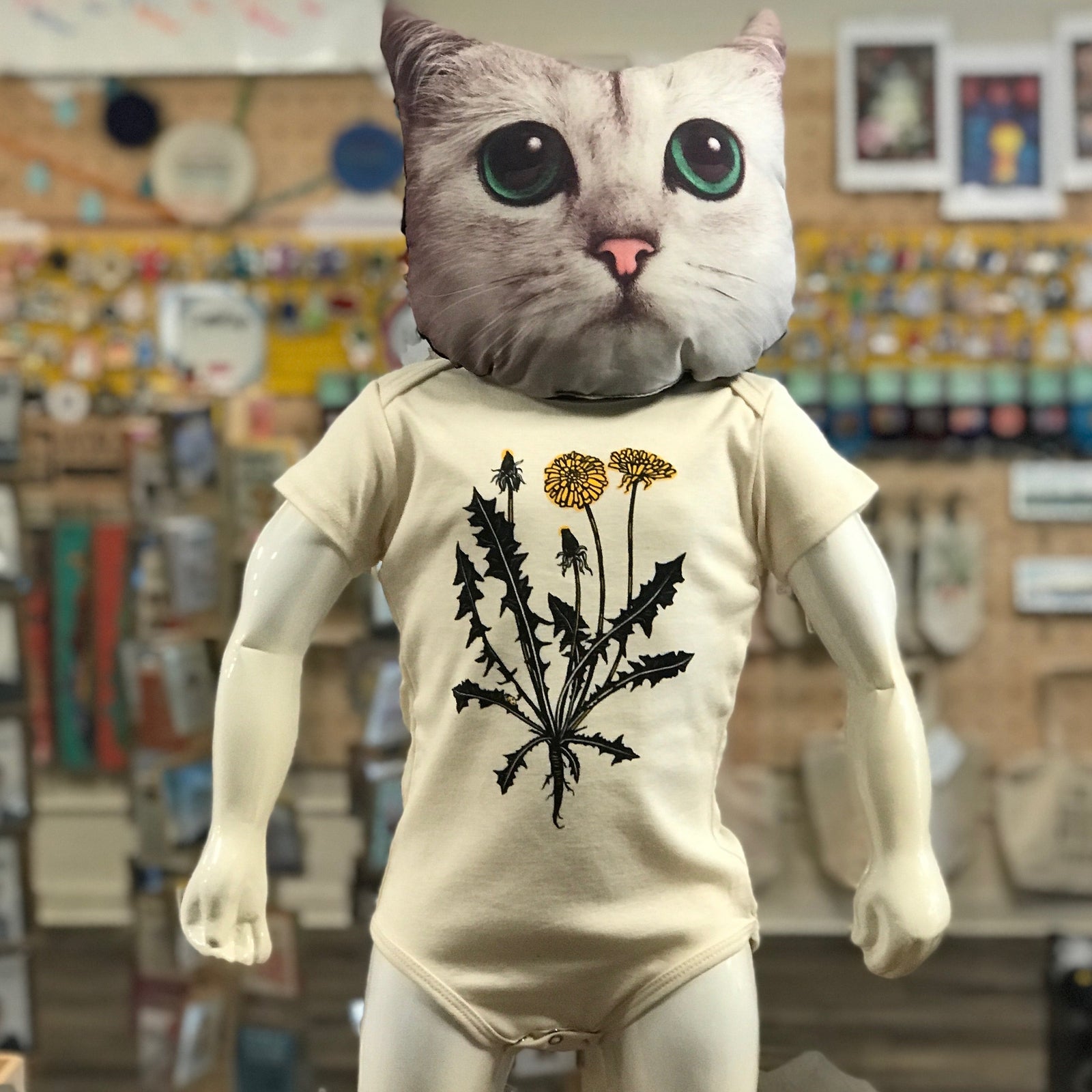 A baby mannequin with a cat head wearing a screenprinted onesie with a dandelion on it. The background shows a blurry store in the background.