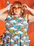 A woman with feathered hair stands in front of an orange background. There is a life preserver and buoys hanging in the background along with a net. She is wearing square sun glasses and a dress covered in a lobster print. The dress has pockets. 