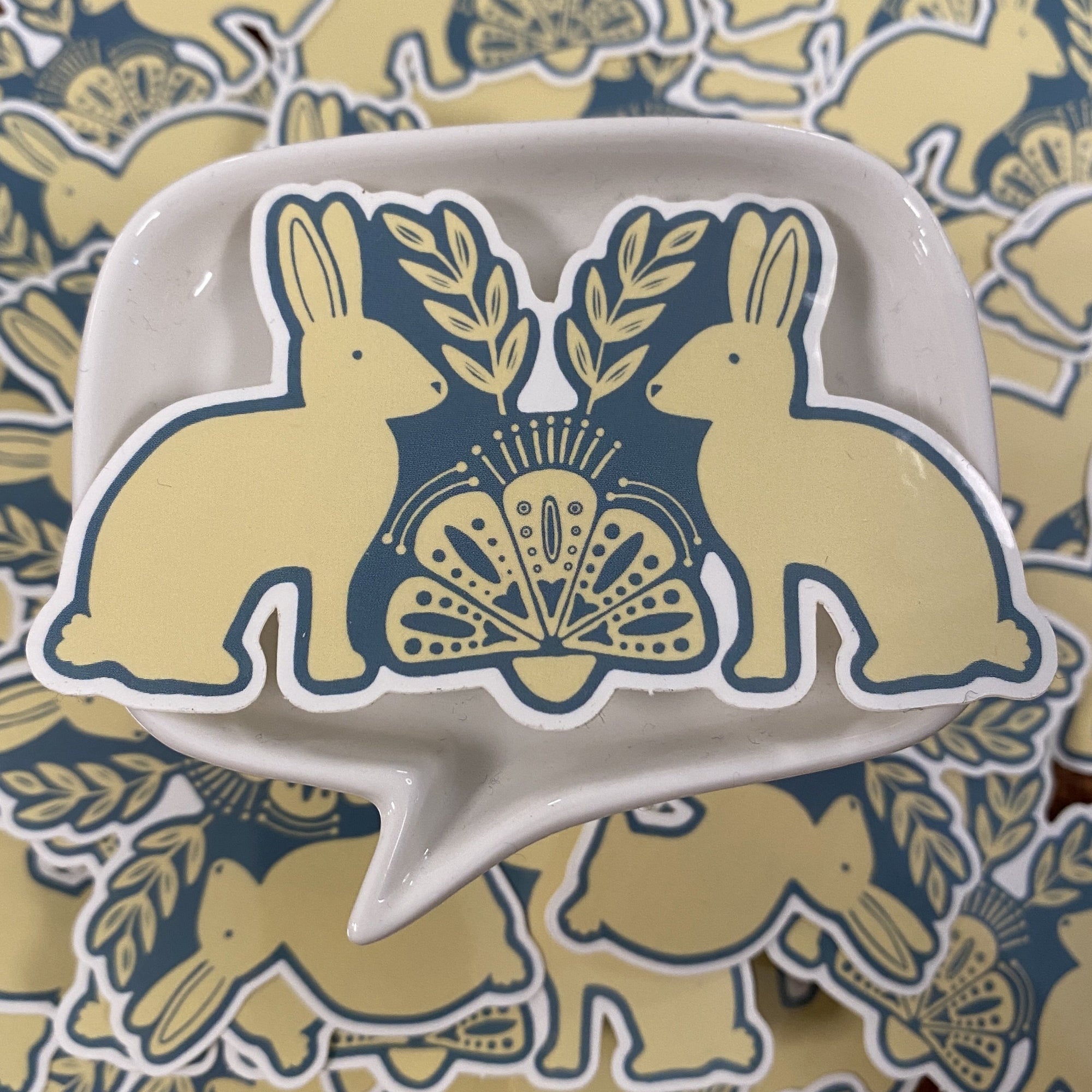 A blue and off-white die cut vinyl sticker by Ugly Baby. It is a folk art design of two rabbits facing each other. There is a folk art design between them.