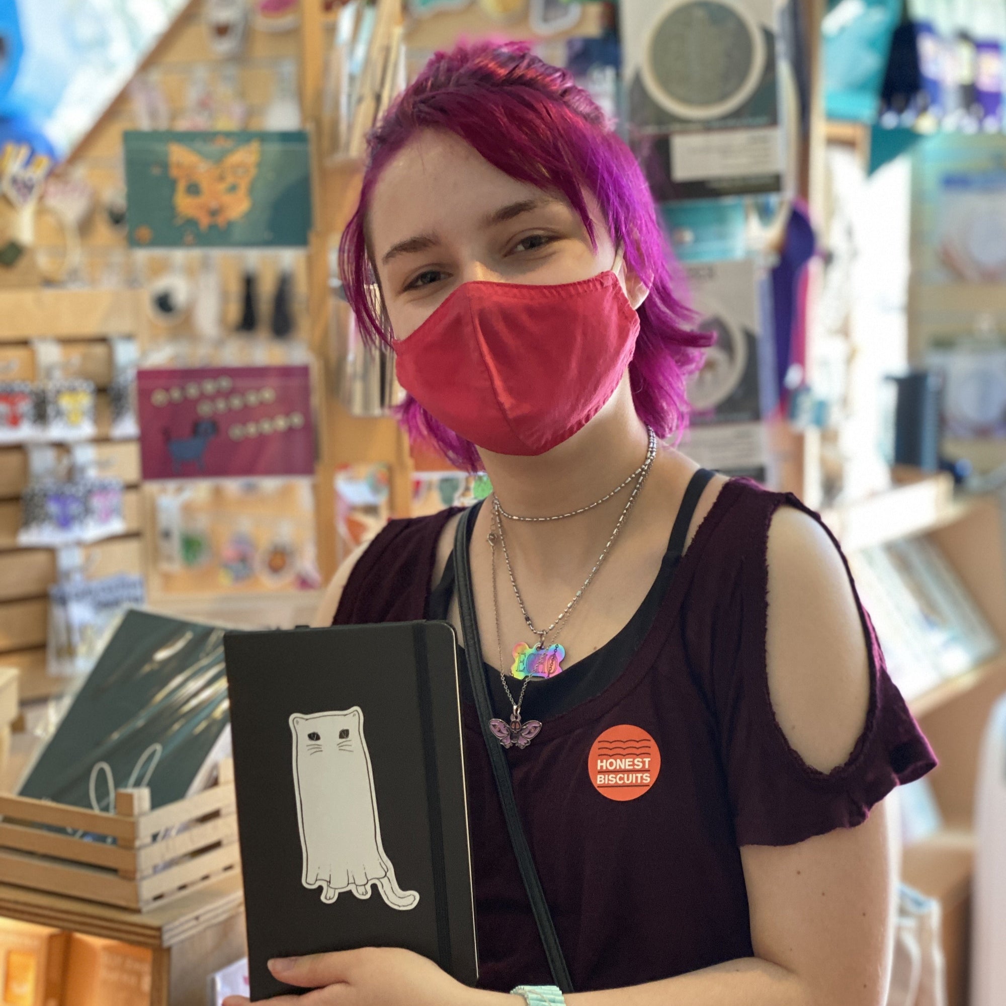A woman with chin-length maroon hair and wearing a red mask is holding a black notebook sporting the Ghost Cat sticker.