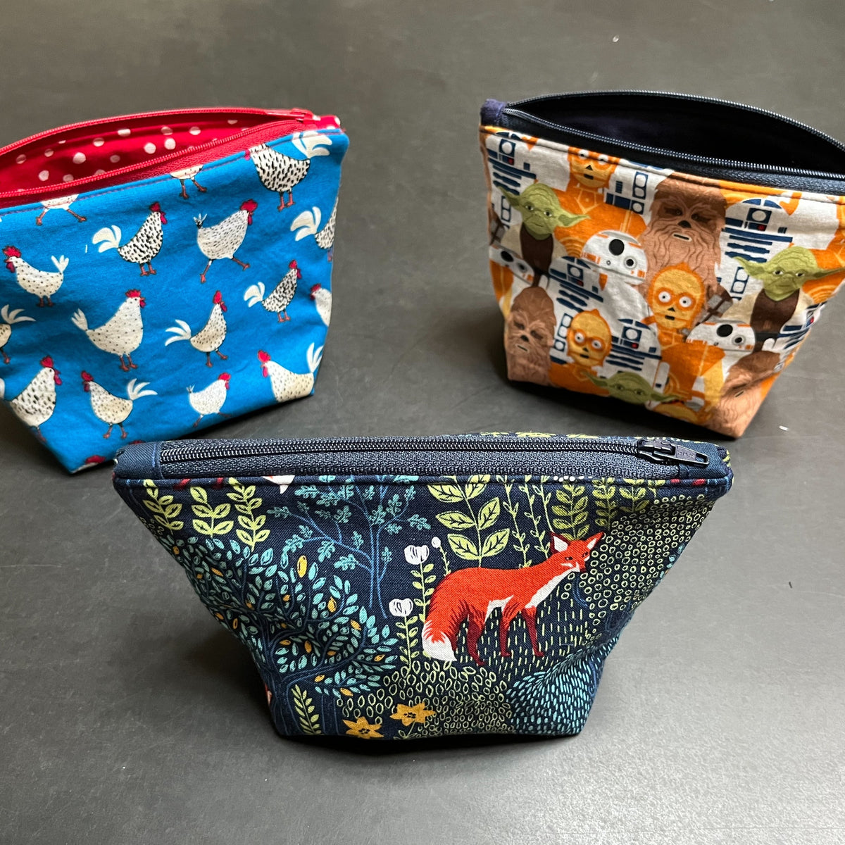 Three examples of zippered box bottom bags. On the left, chickens and roosters on a blue background and lined with red polka dot fabric &amp; a red zipper. On the right, a Star Wars graphic lined with black fabric. In the center front, a woodland theme with pretty green foliage and a red fox. 