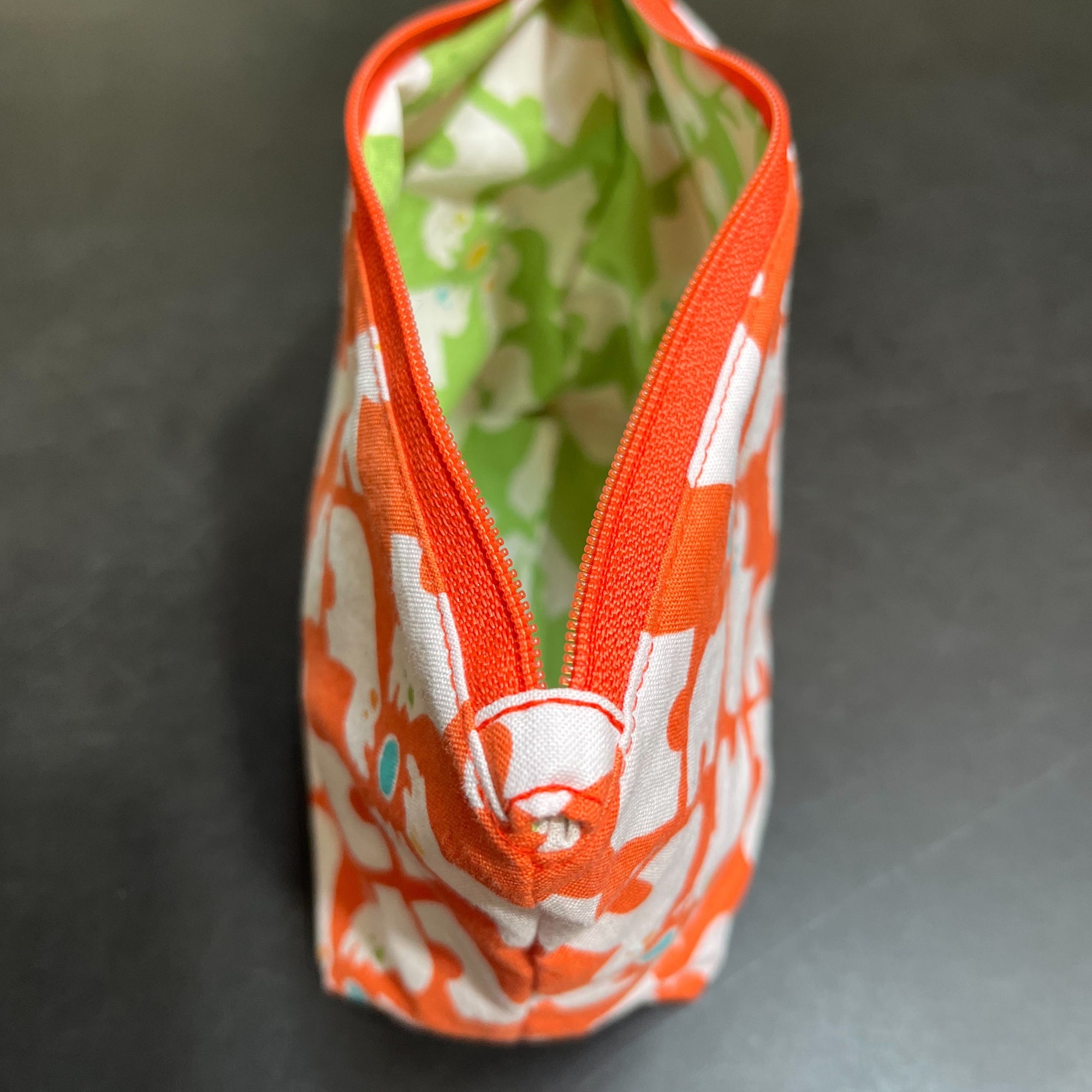 Example of a lined zipper pouch with a box bottom so it stands on its own. The exterior fabric is orange and white and the interior is lime green and white. It has a bright orange zipper. 