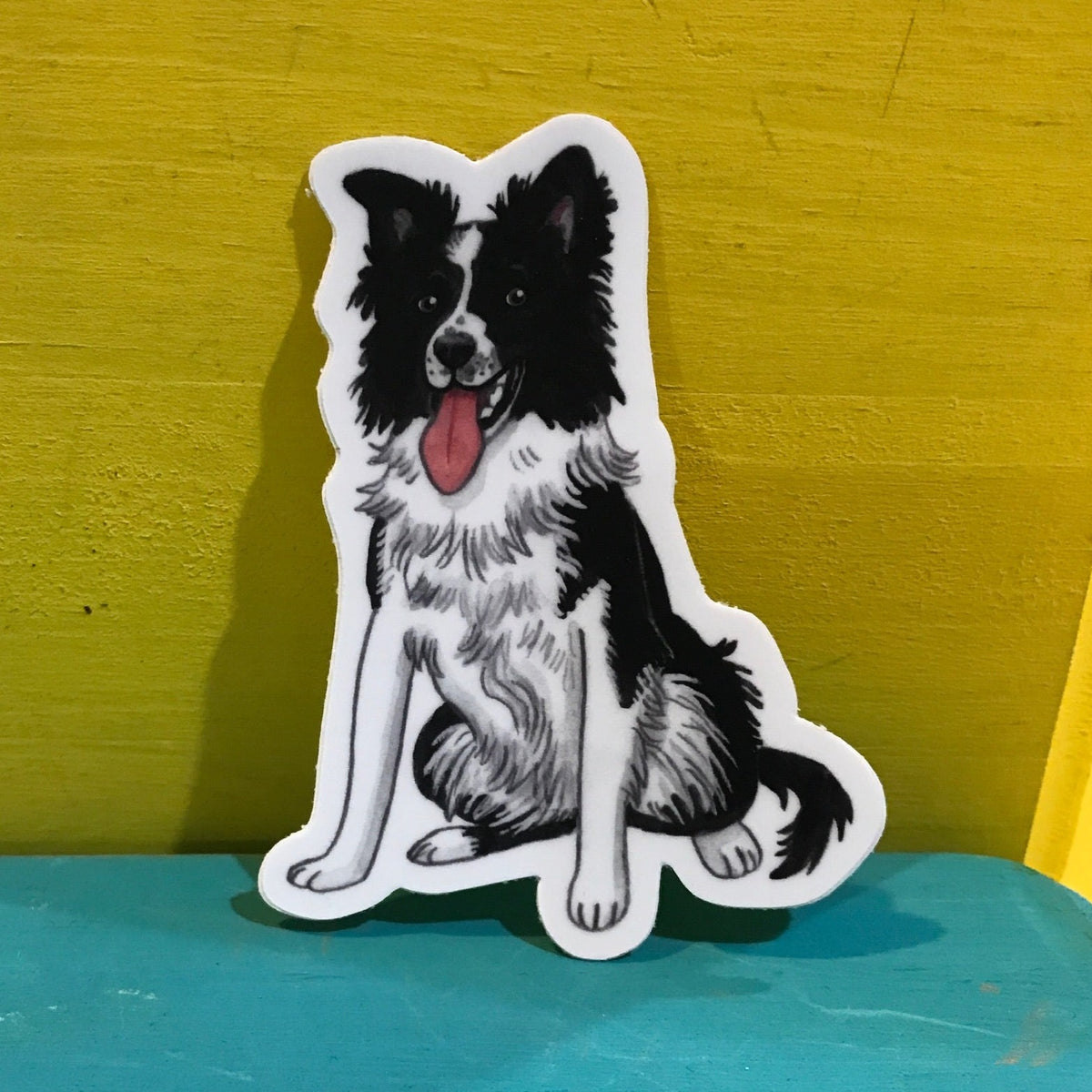 Watercolor illustration of a border collie. It&#39;s a sticker with a white outline and it&#39;s standing up against a yellow and aqua background. 
