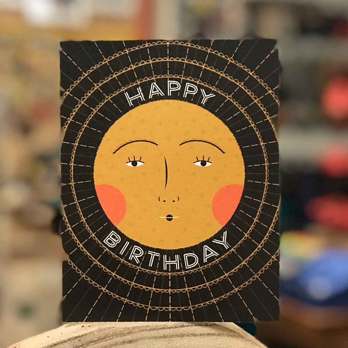 A black card with a mustard yellow sun with rosy cheeks in the middle. It says &quot;Happy Birthday.&quot; The sun haas a pretty face. The card is sitting atop a piece of wood and the background of the photo is blurry. 
