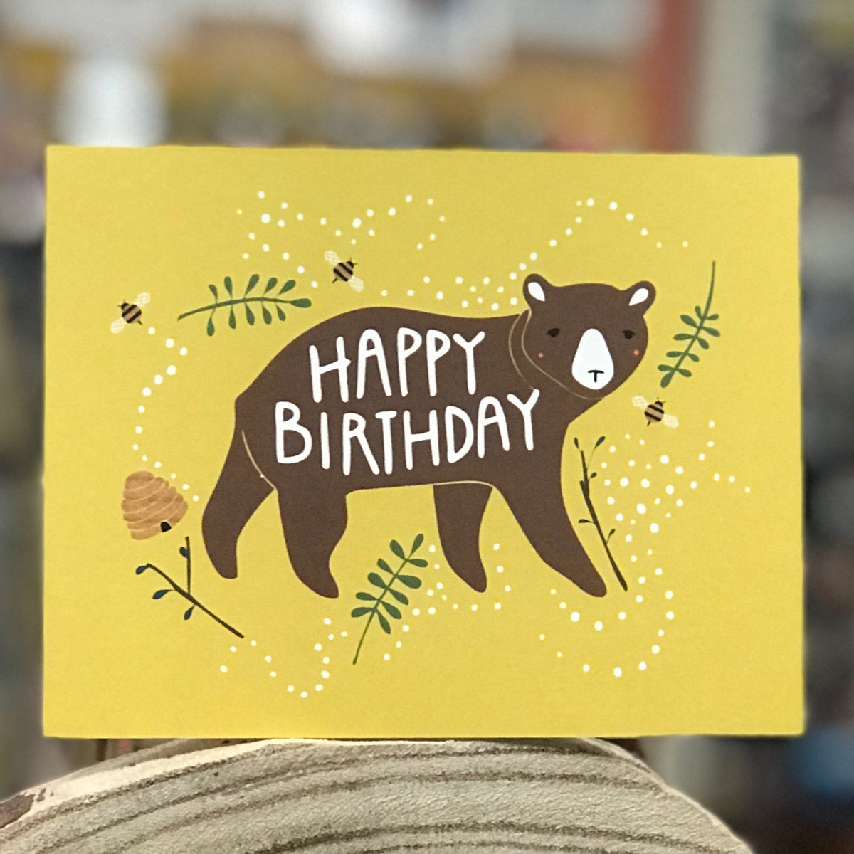 A mustard yellow card shows an image of a brown bear with rosy cheeks. It says &quot;Happy Birthday&quot; across his body. Bees and a hive are in the background along with some greenery. The card is standing on a piece of wood and the background of the shot is blurry. 