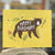 A mustard yellow card shows an image of a brown bear with rosy cheeks. It says "Happy Birthday" across his body. Bees and a hive are in the background along with some greenery. The card is standing on a piece of wood and the background of the shot is blurry. 
