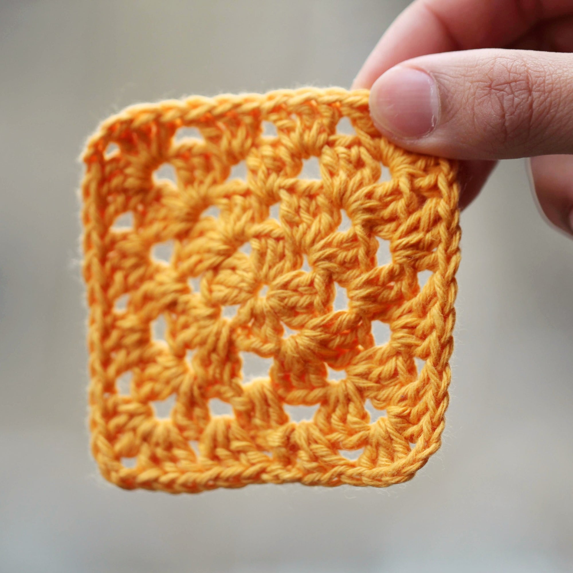  A light orange crocheted granny square being held up by a hand against a blurry background.