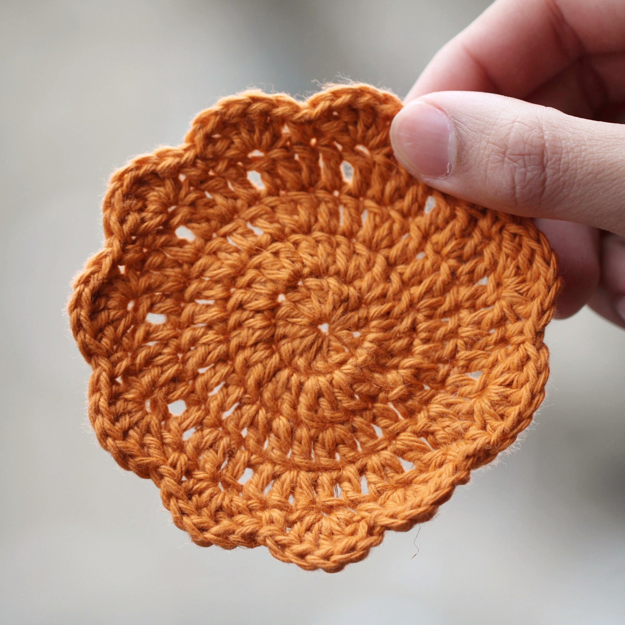 A round crocheted coaster with a scalloped edge. It is light orange and being held up my a human hand against a blurry background.