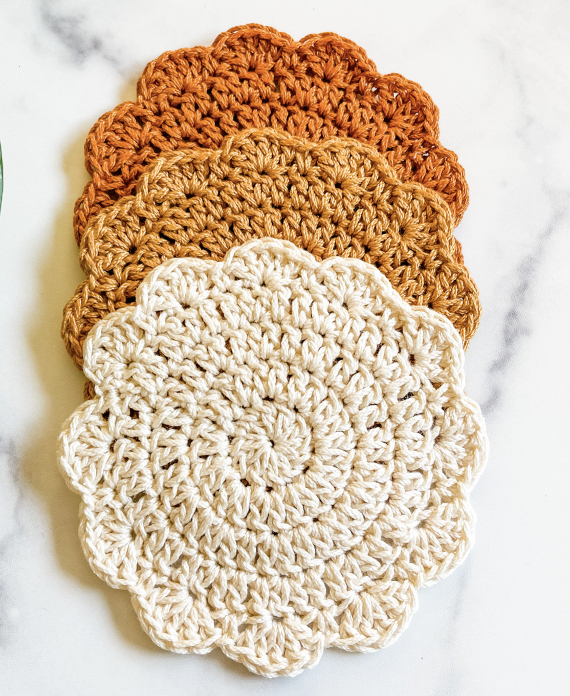 A set of three crocheted coasters with a scalloped edges, They are dark orange, light orange and off white. They are sitting on a marble tabletop. 
