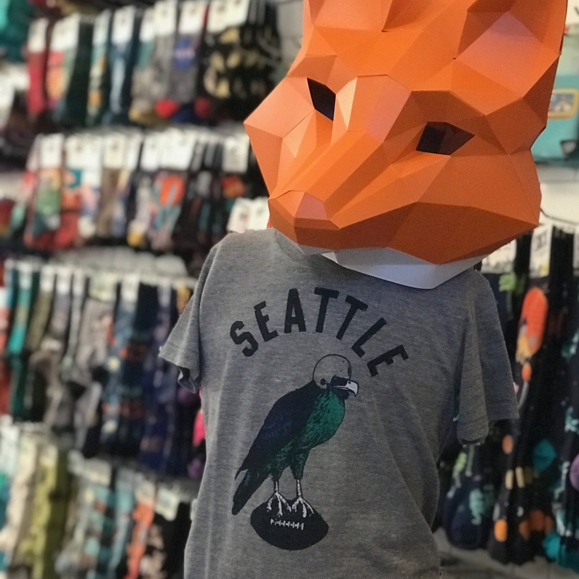 A heather gray shirt that says SEATTLE in block letters. Below there is a seahawk wearing a helmet and standing on a football. It is shown on a mannequin wearing an orange fox mask made of paper. In the background there is a wall of socks blurred. 