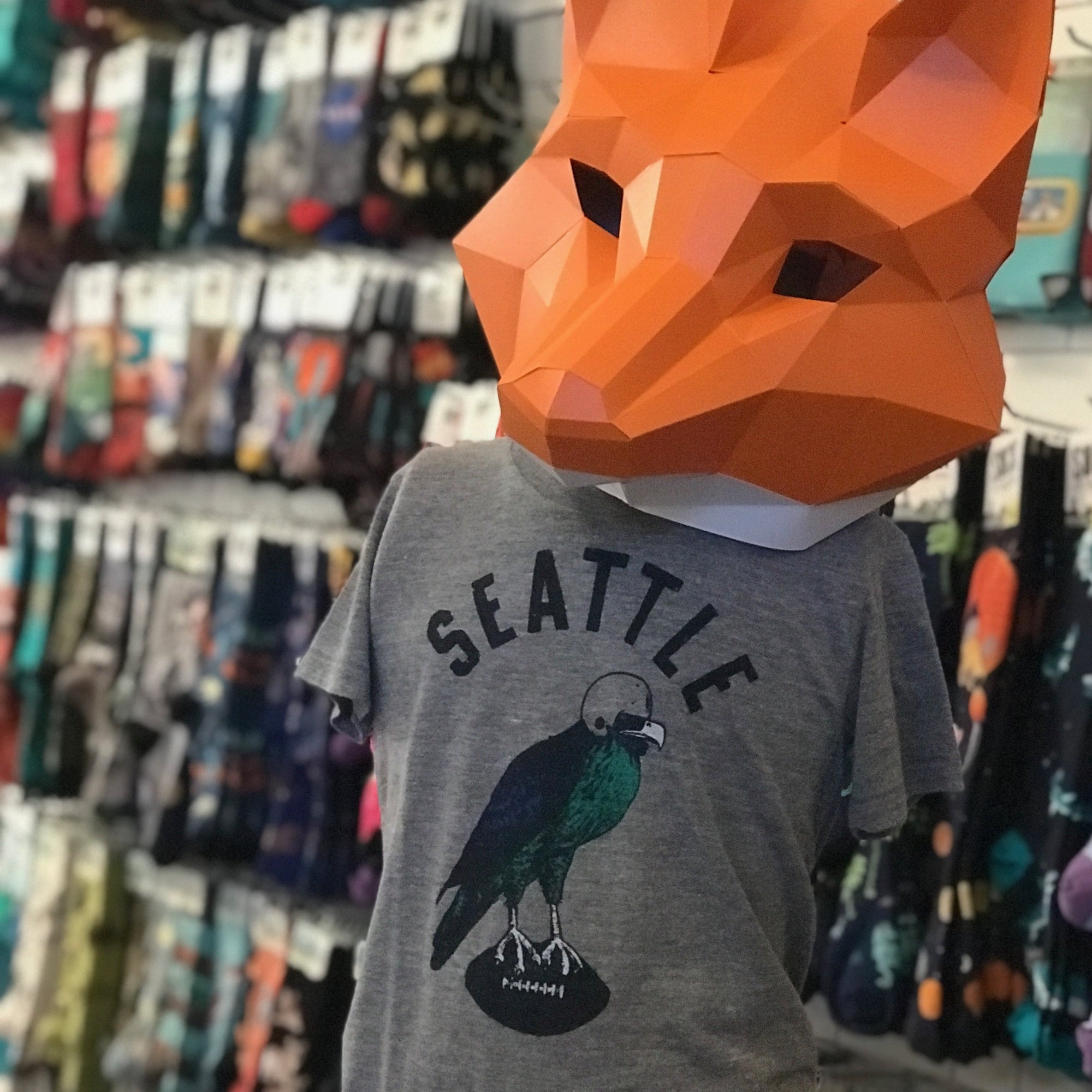 A heather gray shirt that says SEATTLE in block letters. Below there is a seahawk wearing a helmet and standing on a football. It is shown on a mannequin wearing an orange fox mask made of paper. In the background there is a wall of socks blurred.
