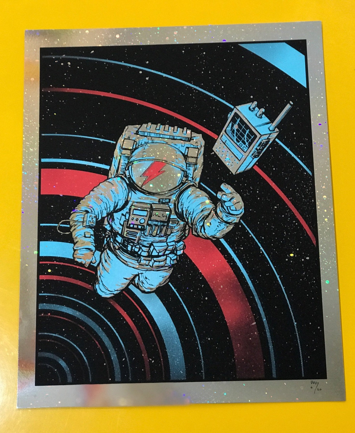 An astronaut screenprinted with holographic accents. The astronaut has a David Bowie red lightning bolt on the helmet. The astronaut is reaching out for a communication device. They are floating in space with red and blue circular lines surrounding them in the background.