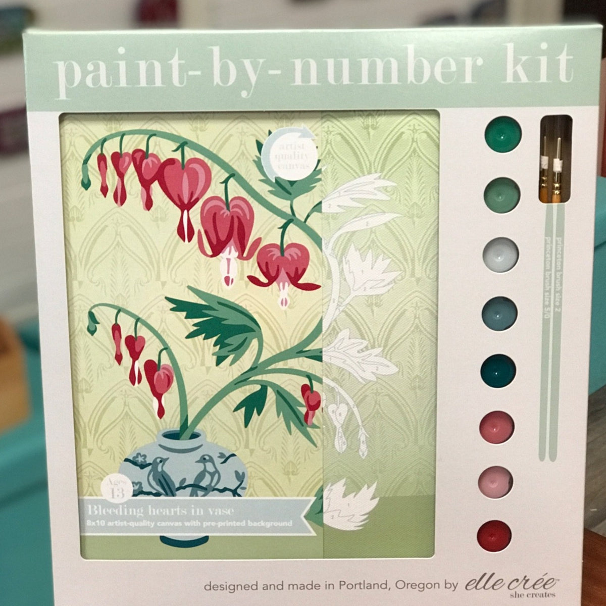 Paint by number kit package with image of bleeding hearts plant on the left and paint colors on the right. The flowers look like hearts and a stem of them are in a blue vase with birds on itl