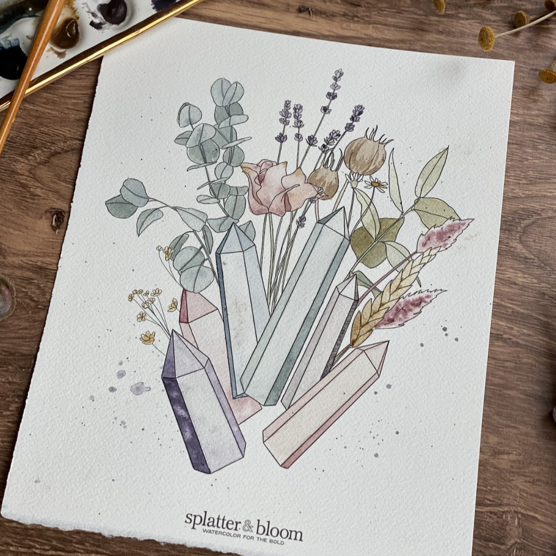 A watercolor painting of crystals and flowers in muted tones. A watercolor paint set and paint brush is in the upper left corner. The background is a wood grain table. 