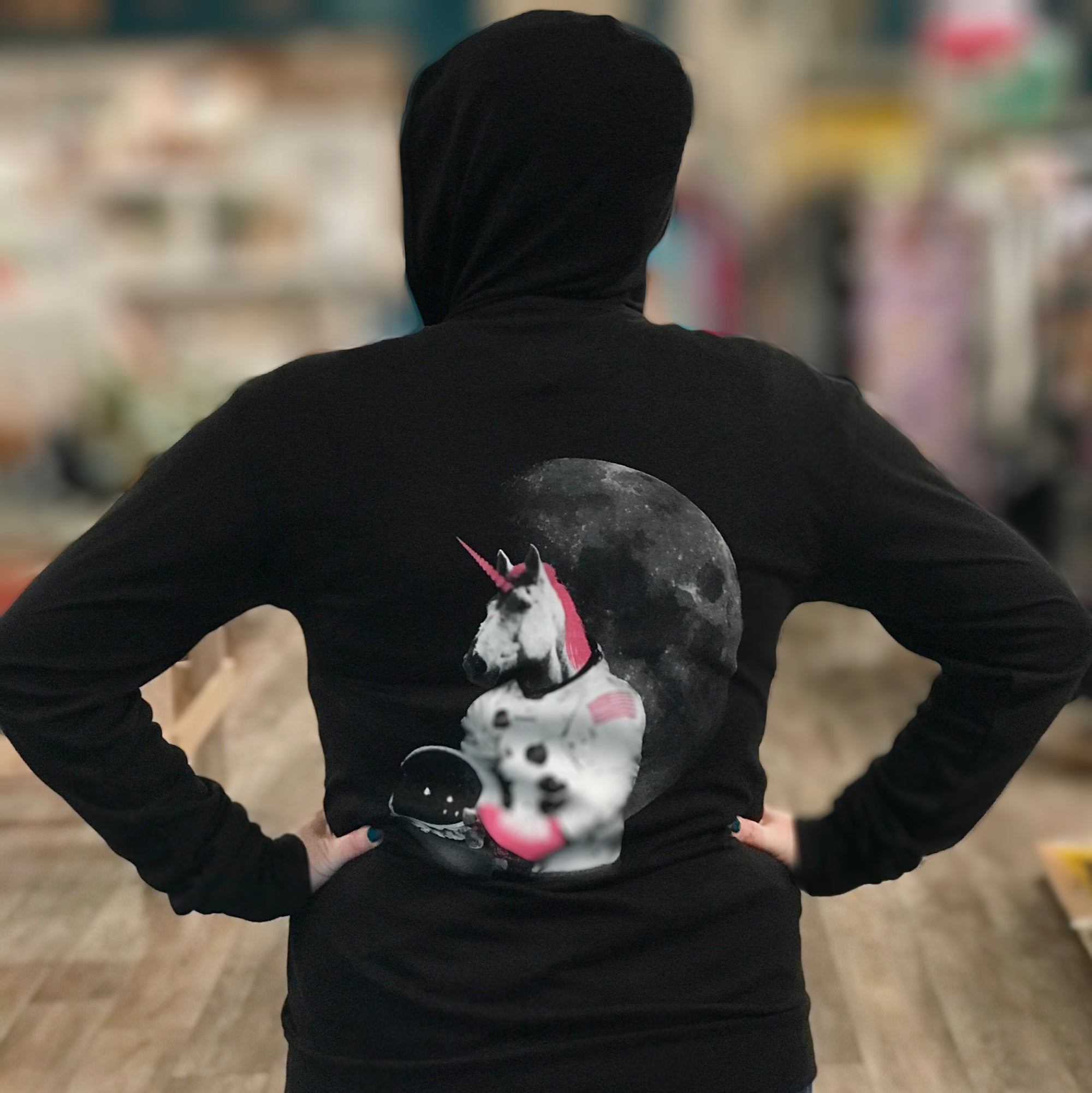 Black hooded sweatshirt with a roller skating unicorn astronaut screenprinted on the back in white and hot pink.  A person models with the hood up and their hands on their hips. 