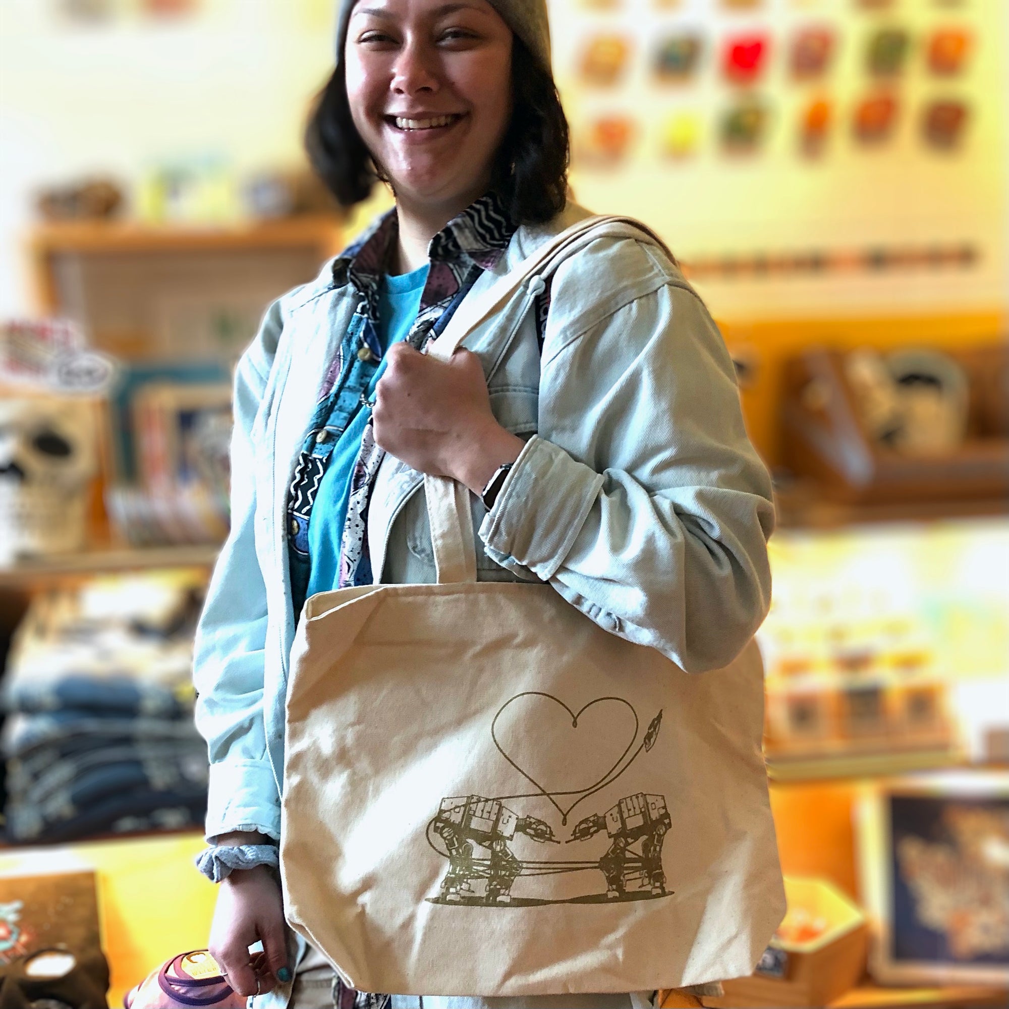 A smiling dark haired woman wearing a gray beanie is holding an off white color tote bag with two AT-ATs falling in love.